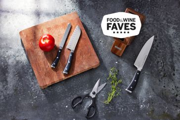 Three knifes on a counter with cutting board, scissors and tomato with food and wine badge in top right.