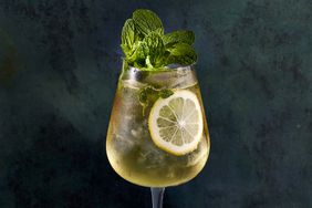 Close up of a Hugo Spritz cocktail in a wine glass, pale yellow with a lemon wheel against the side of the glass and a large bouquet of mint