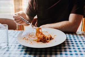 A man sits at a table, covered with a blue checkered tablecloth. He uses a fork to scoop up a mouthful of long spaghetti pasta. 