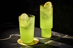 Two neon-green Midori Sour highballs in tall glasses with lemon wheel garnishes, on black background